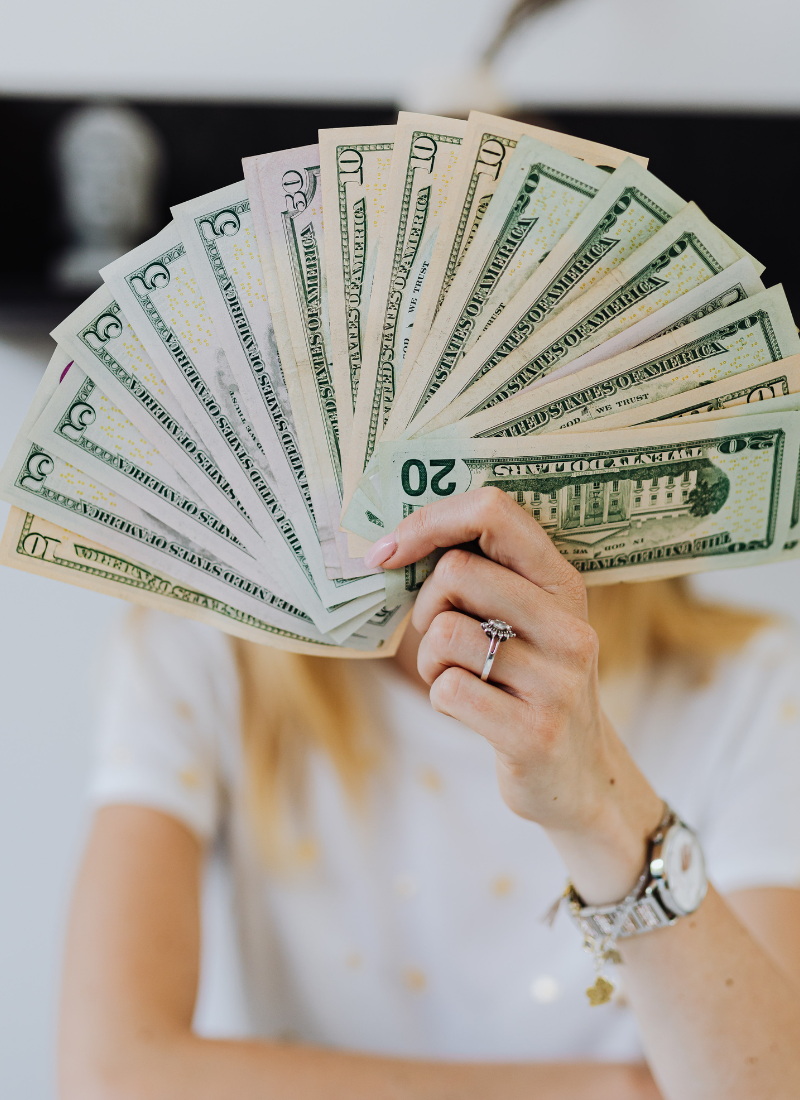 Image of a Women Holding Cash that is Fanned out in front of her Face. The imae leads to the Manifesting Money Category of the blog.